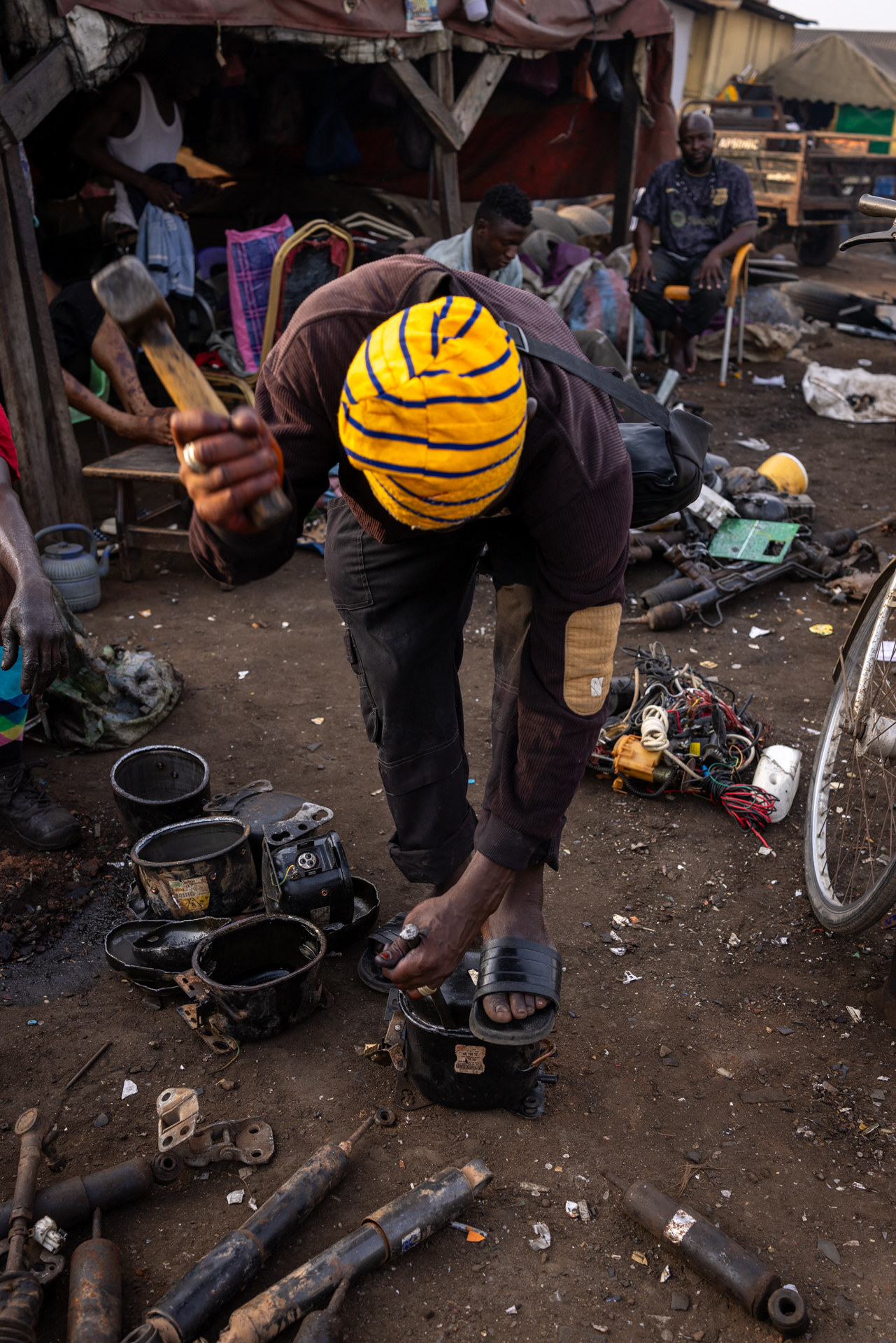 Worker dismantling a compressor with a hammer, e-waste components scattered on the ground